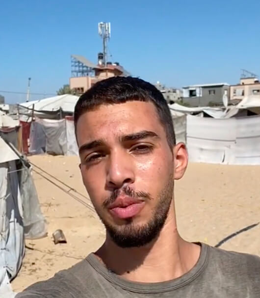 Selfie von Mohammed Said al-Halimy: Ein junger Mann blickt in die Kamera, hinter ihm weiße Zelte auf sandigem Boden.