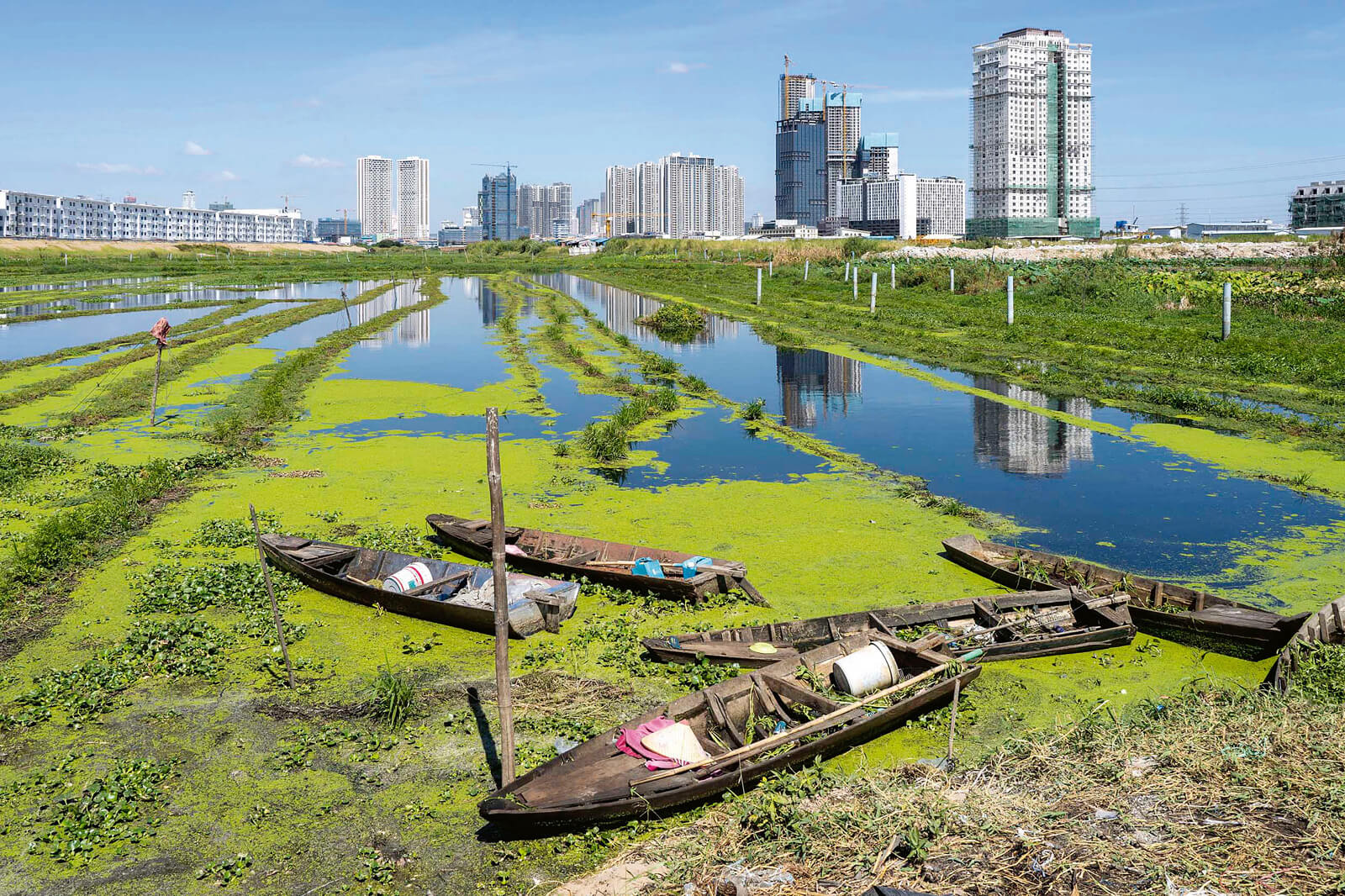 Fotografie eines Gewässers mit kleinen Booten darauf, im Hintergrund moderne Wolkenkratzer.