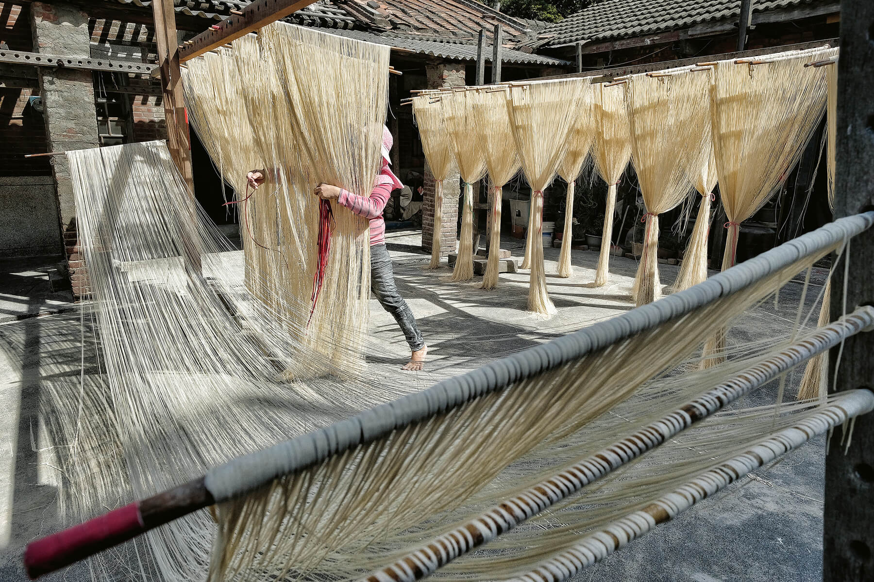 Eine Frau mit Hut bindet lange Nudeln zusammen, die in einem Hof zum Trocknen hängen.