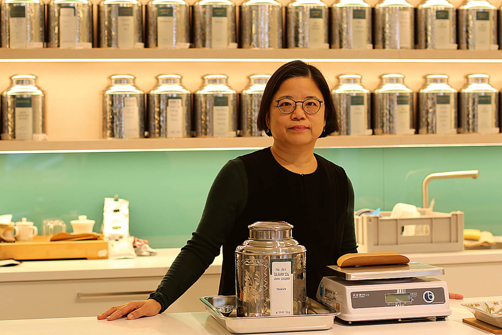 Porträt von Feng-Yu Hsieh: Eine Frau mit Brille steht hinter der Theke eines Teeladens, im Hintergrund zahlreiche Teebehälter.