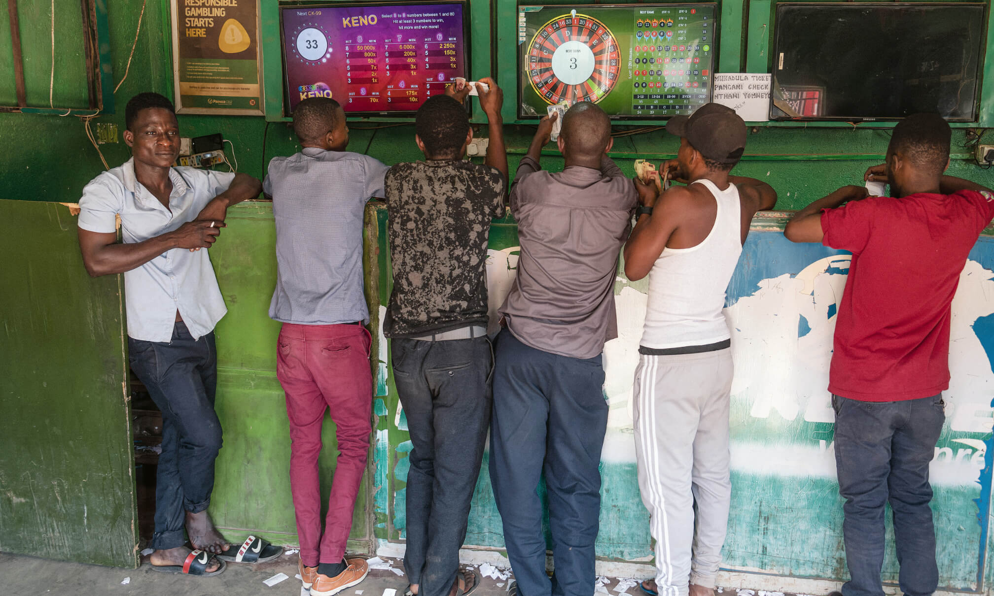 Mehrere Männer beim Wetten, alle haben Bargeld in der Hand, alle bis auf einen blicken in Richtung mehrerer Bildschirme, auf denen digitales Glücksspiel zu verfolgen ist.