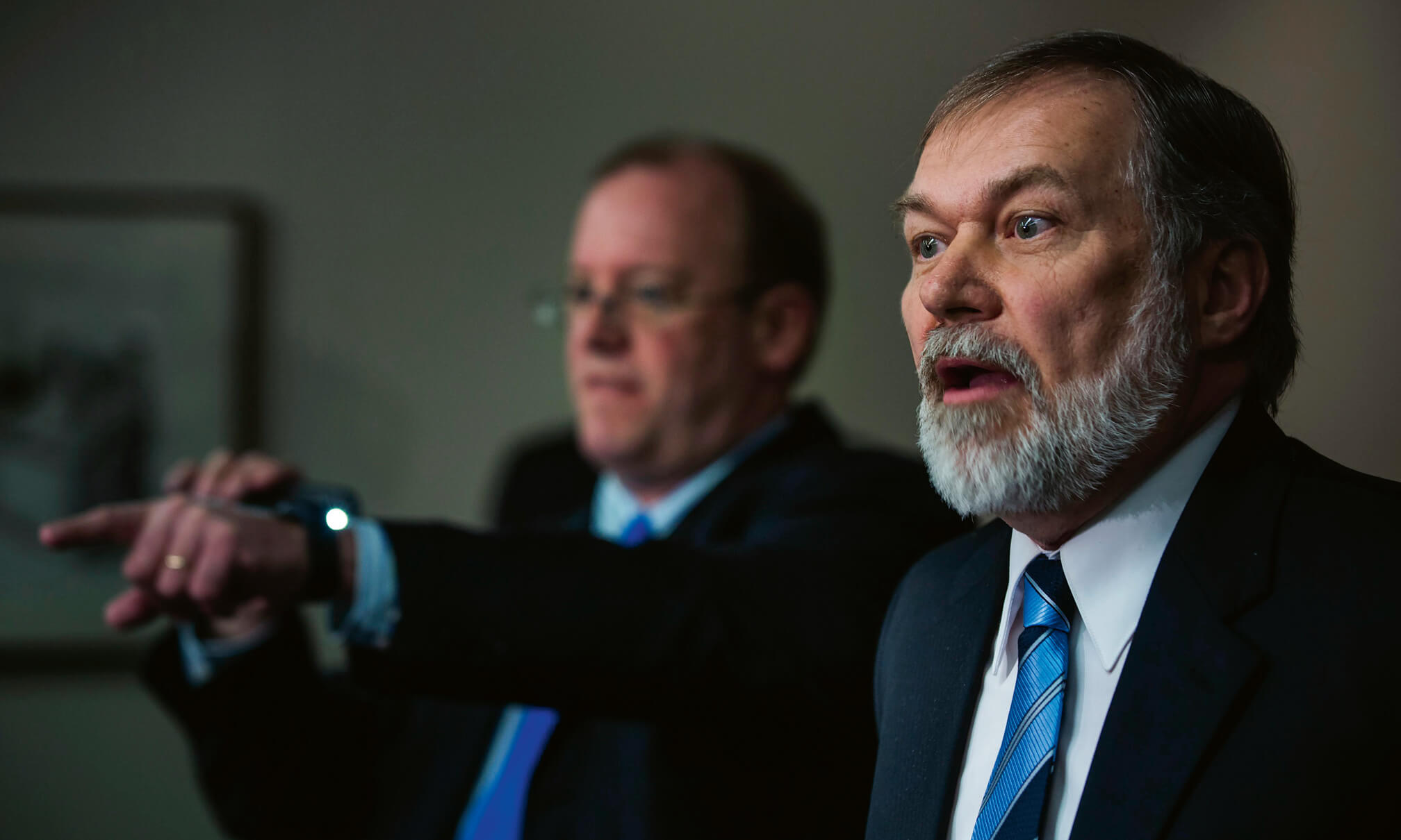 Zwei Männer in Anzug, der Mann im Hintergrund deutet auf etwas, jener im Vordergrund scheint aufgebracht zu sein.