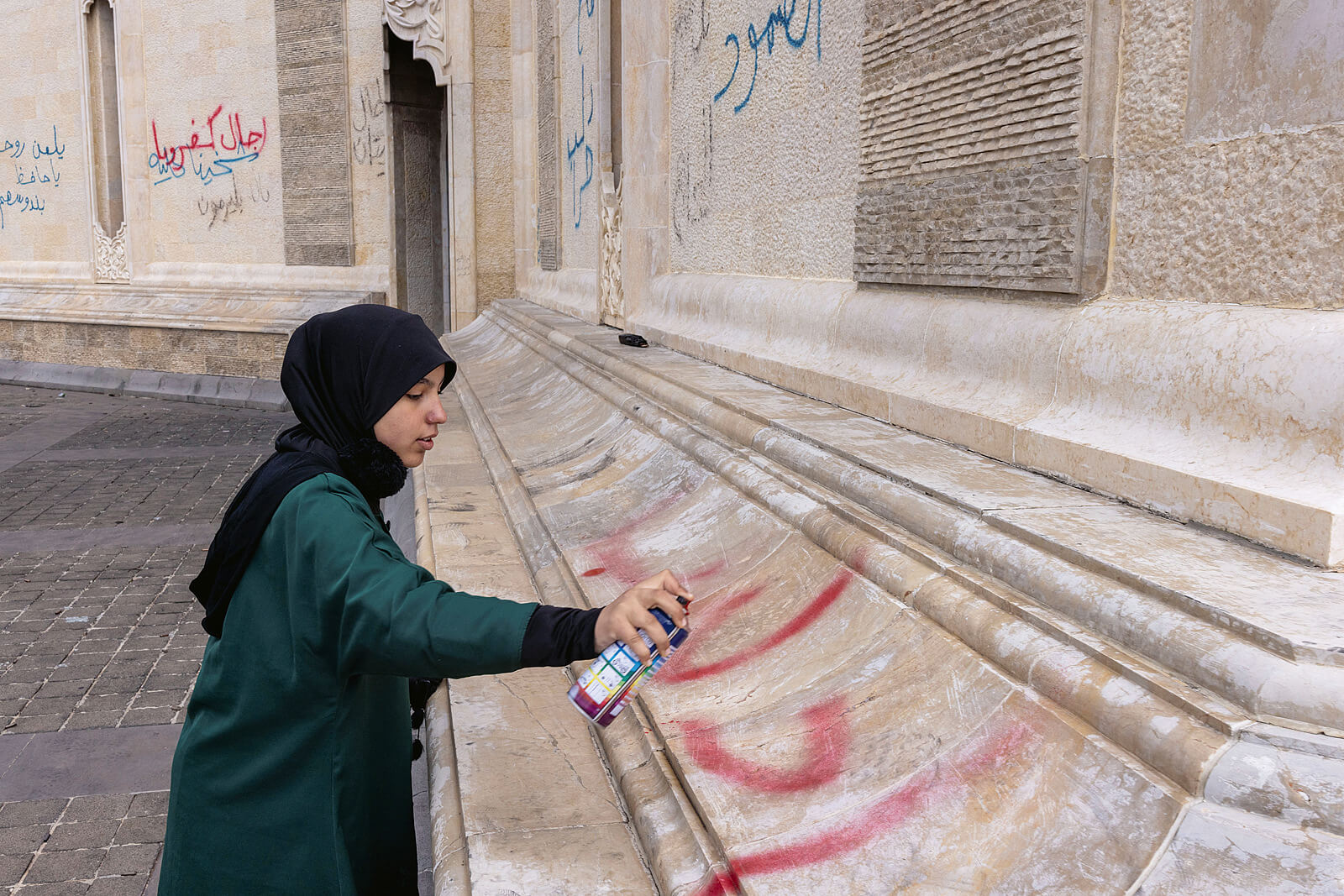 Eine junge Frau mit schwarzem Kopfuch und eine grünen Kleid sprüht einen Graffiti-Schriftzug in Rot an eine sandsteinfarbene Fassade.