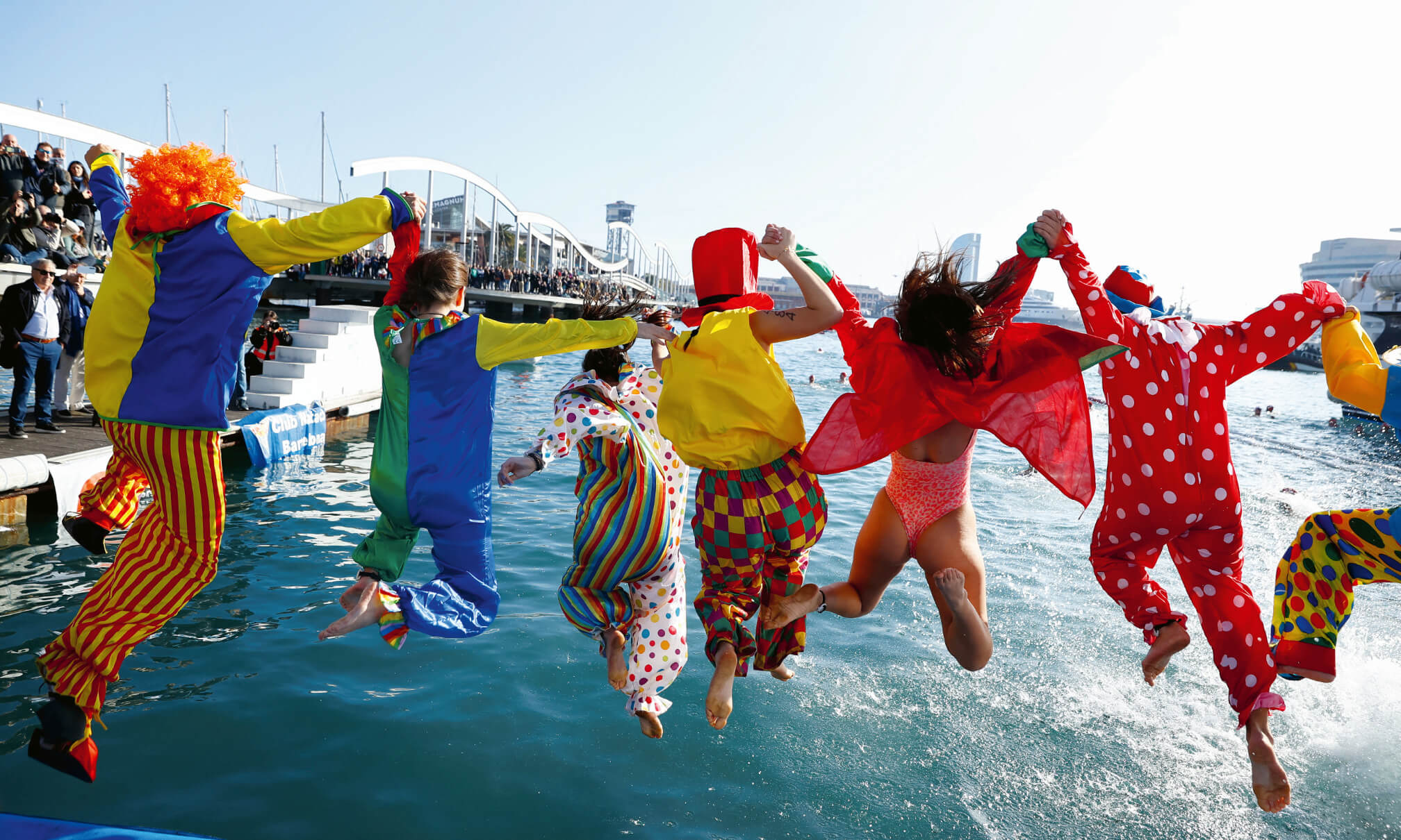 Eine Gruppe von als Clowns verkleideten Menschen springt ein ein Gewässer, die Personen halten sich dabei an den Händen.
