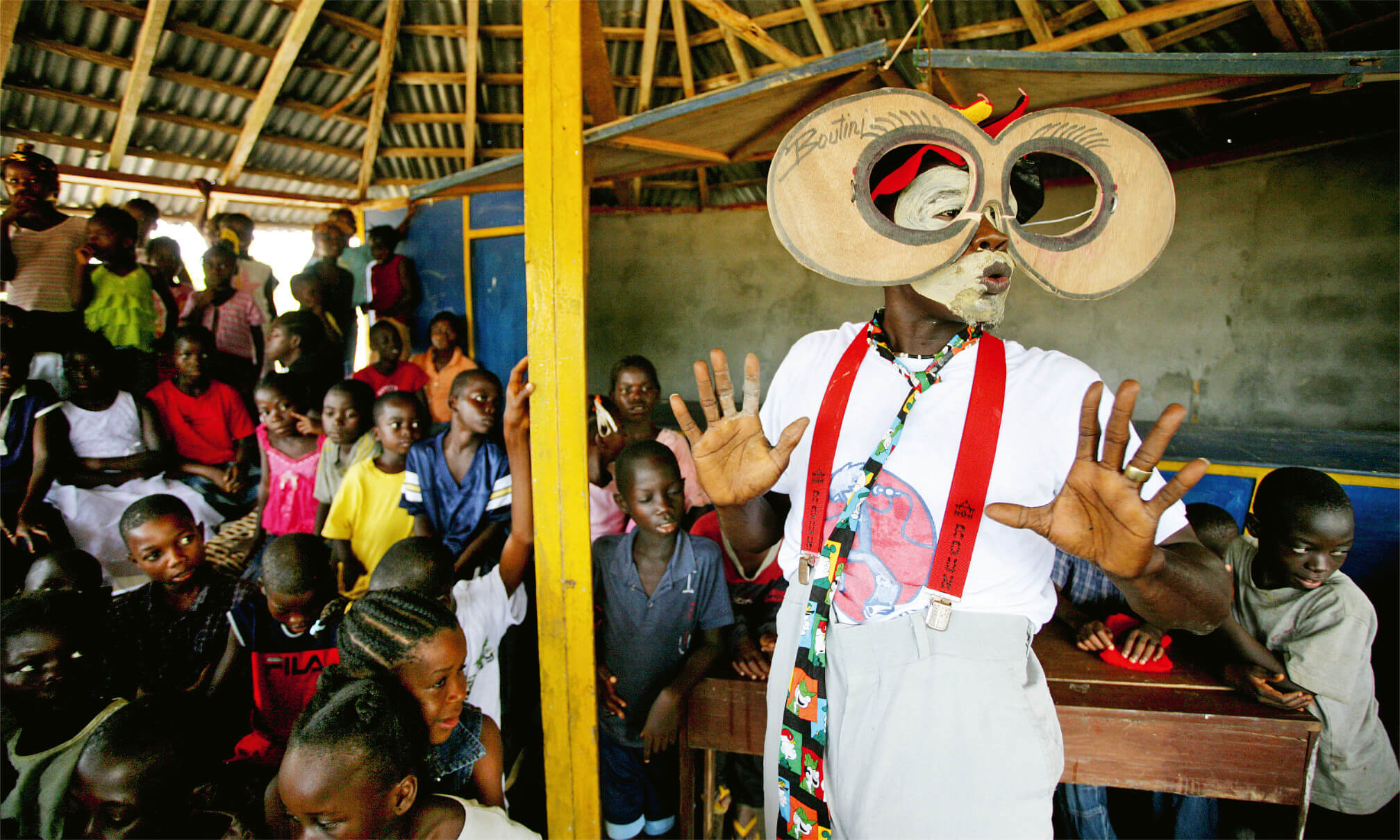 Ein Mann mit großer, runder Brille und bunter Kleidung unterhält eine Gruppe von Kindern in einem einfachen Klassenraum mit Holzdach.