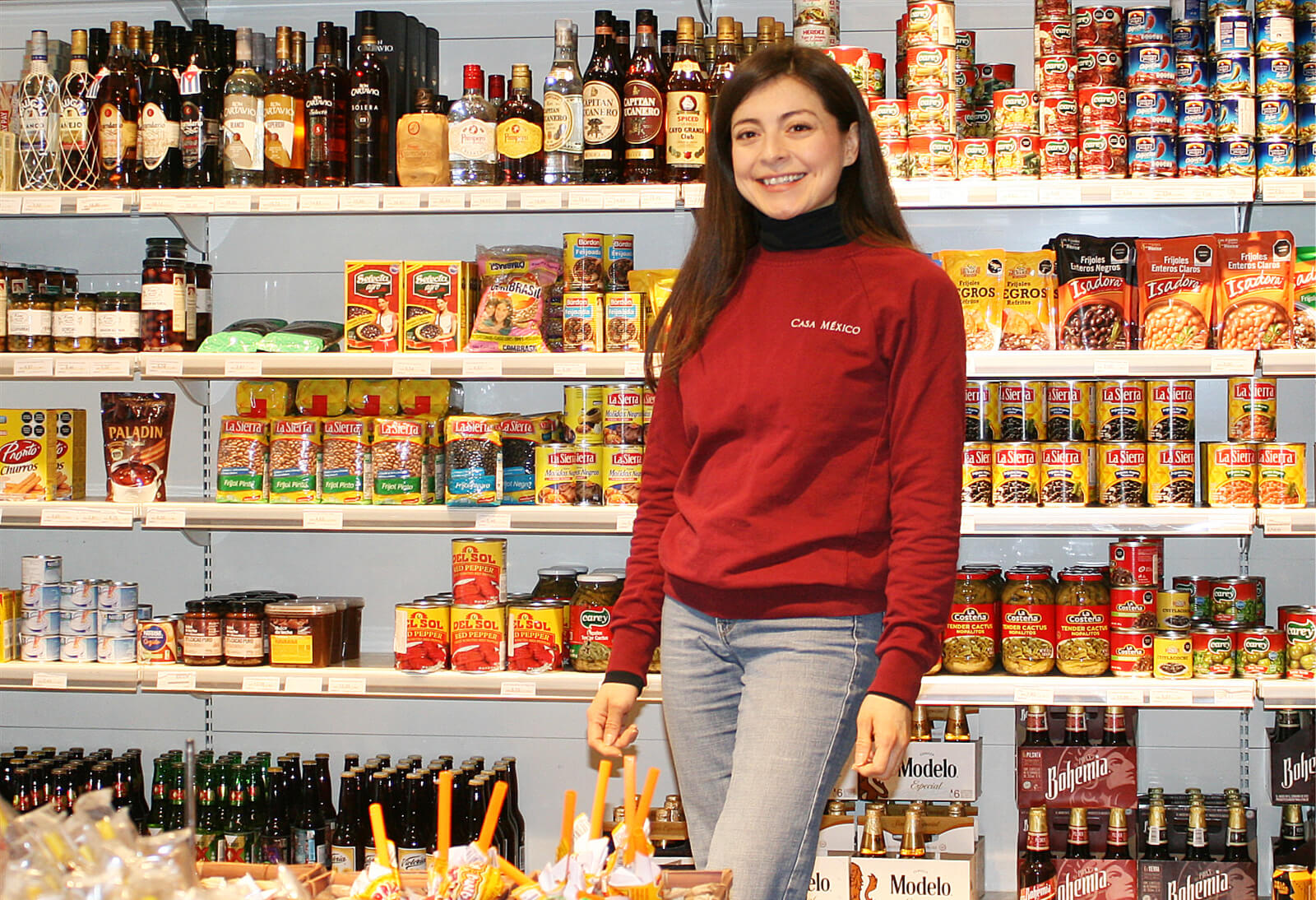 Porträt von Iliana Márquez: Eine Frau steht vor einem gut gefüllten Regal in einem Lebensmittelgeschäft und lächelt.