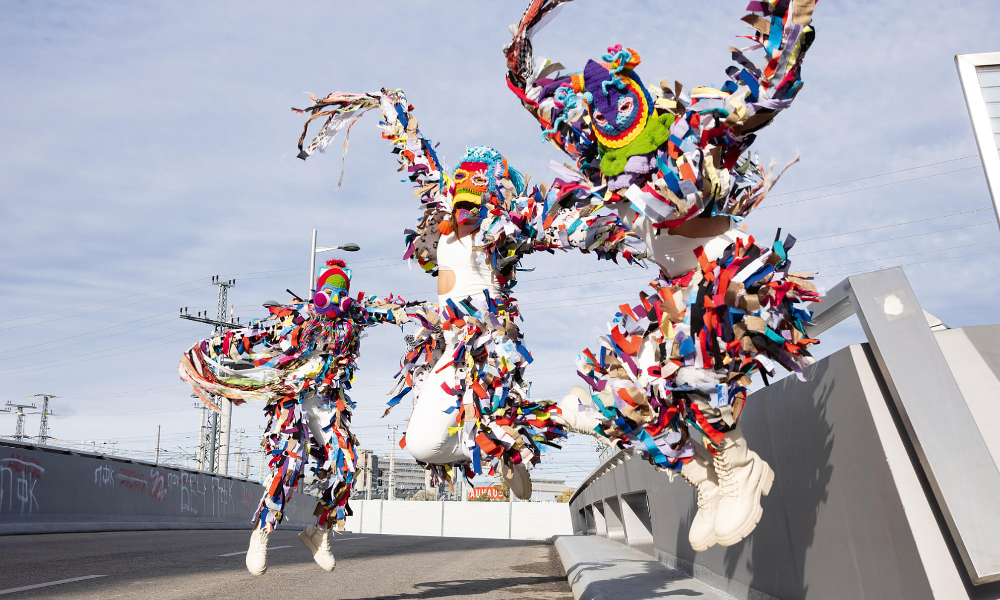 Drei Menschen in bunten, fransigen Kostümen springen energiegeladen in die Luft – Straßenszene mit kreativem, urbanem Flair.