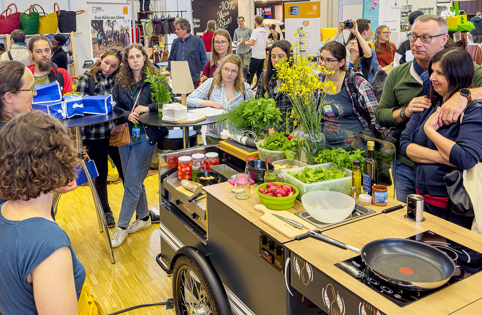 Viele Menschen, die um eine mobile Küche mit Kochutensilien und Zutaten stehen, sie lauschen den Worten einer Person.