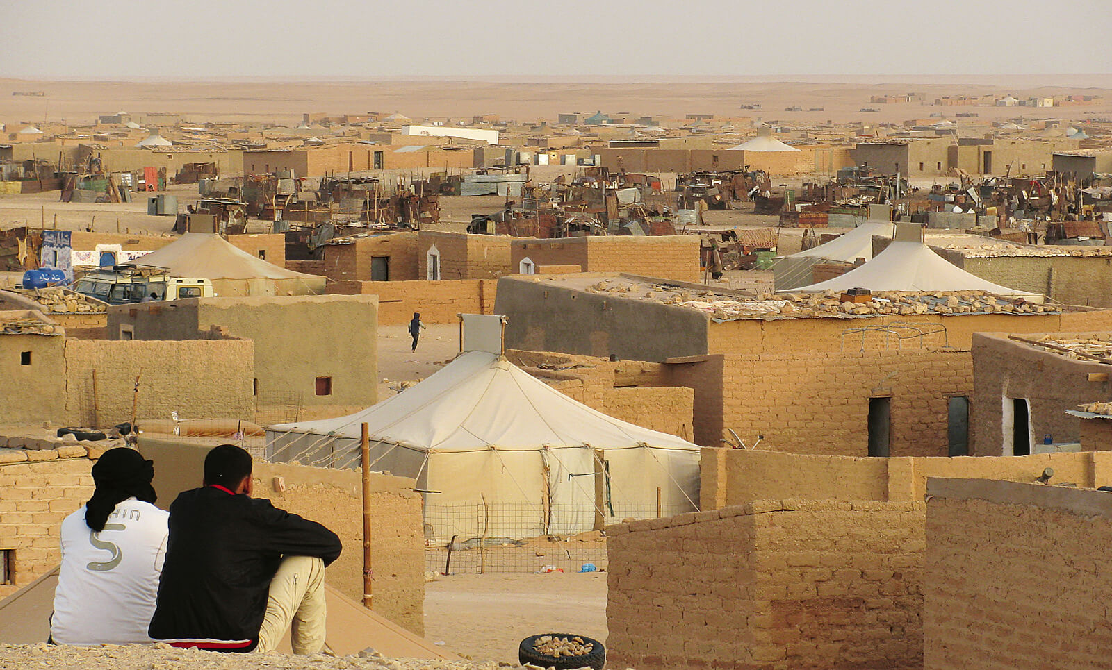 Ansicht eines sahrauischen Flüchtlingscamps in der Westsahara: Zwei Männer blicken ertwas erhöht sitzend auf eine Landschaft aus hellen Zelten und einfachen sandsteinfarbenen Gebäuden in der Wüste.