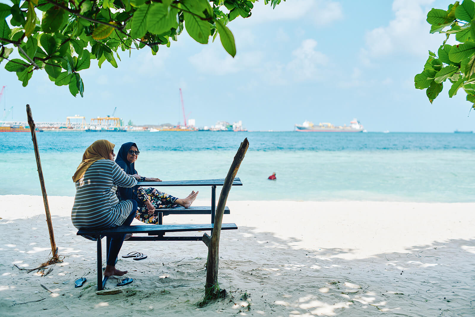 Zwei Frauen mit Kopftuch, die an einem weißen Sandstrand auf je einer Bank an einem Tisch sitzen, im Hintergrund blaues Meer und Schiffe.