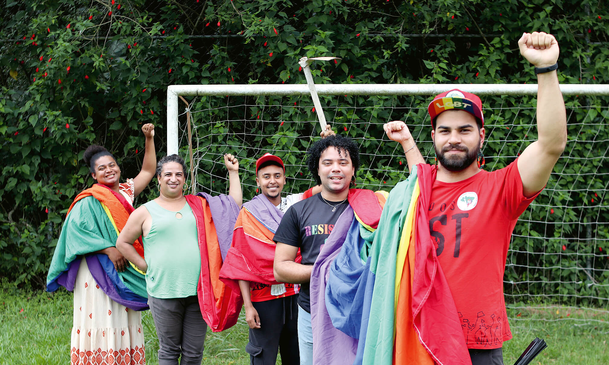 Eine Gruppe von fünf Personen in bunter Kleidung, die alle die linke Faust in die Höhe halten, im Hintergrund ein Fußballtor und grüne Büsche.