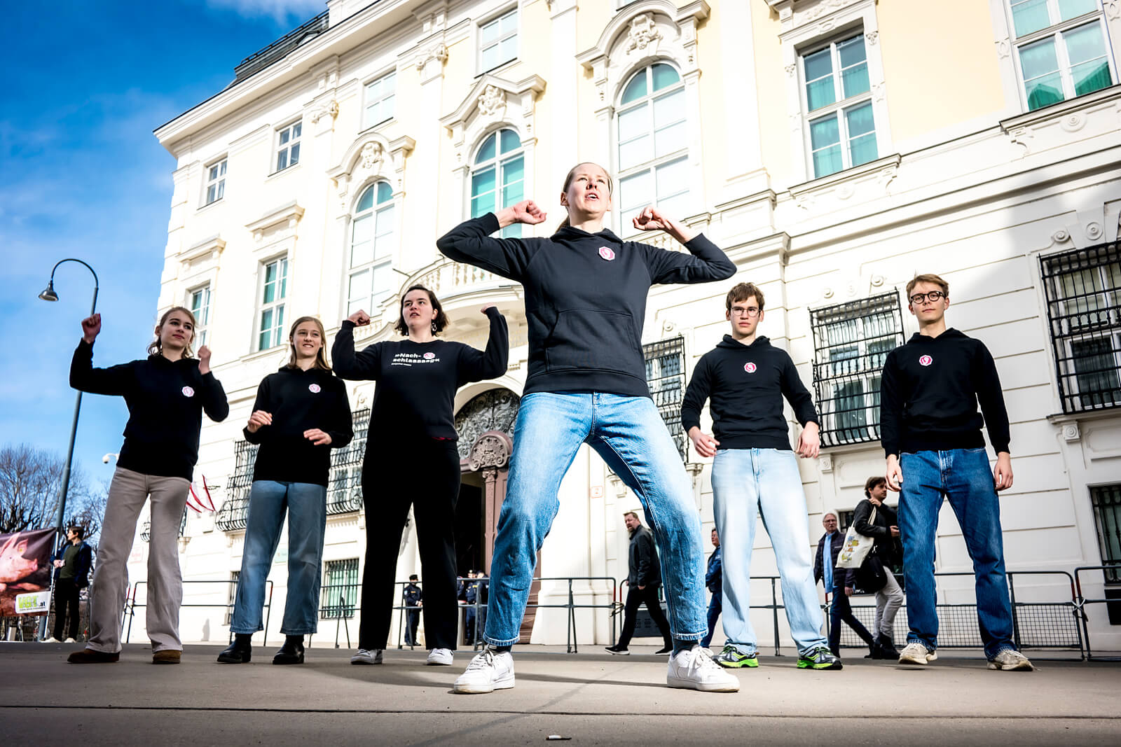 Der Blick von leicht unten auf eine Gruppe von sechs Menschen, die vor einem hellen Gebäude stehen und tanzen.