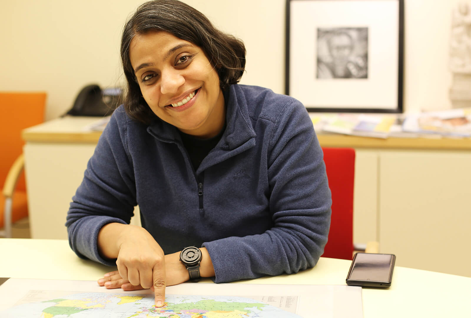 Porträt von Neha Arora: Eine Frau lächelt freundlich in die Kamera, sie deutet mit ihrem rechten Zeigefinger auf eine Landkarte, die vor ihr auf einem Tisch liegt.