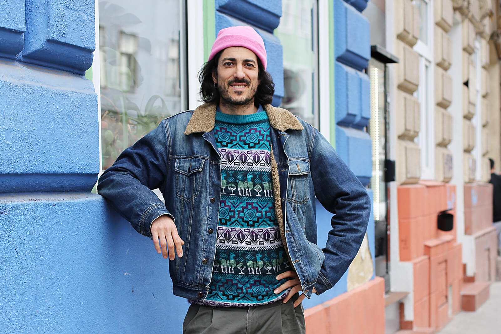Porträt von Nicolás Ramallo: Der Mann steht lässig draußen angelehnt vor einer blauen Häuserwand, er trägt eine rosafarbene Mütze, eine Jeansjacke und lächelt leicht.