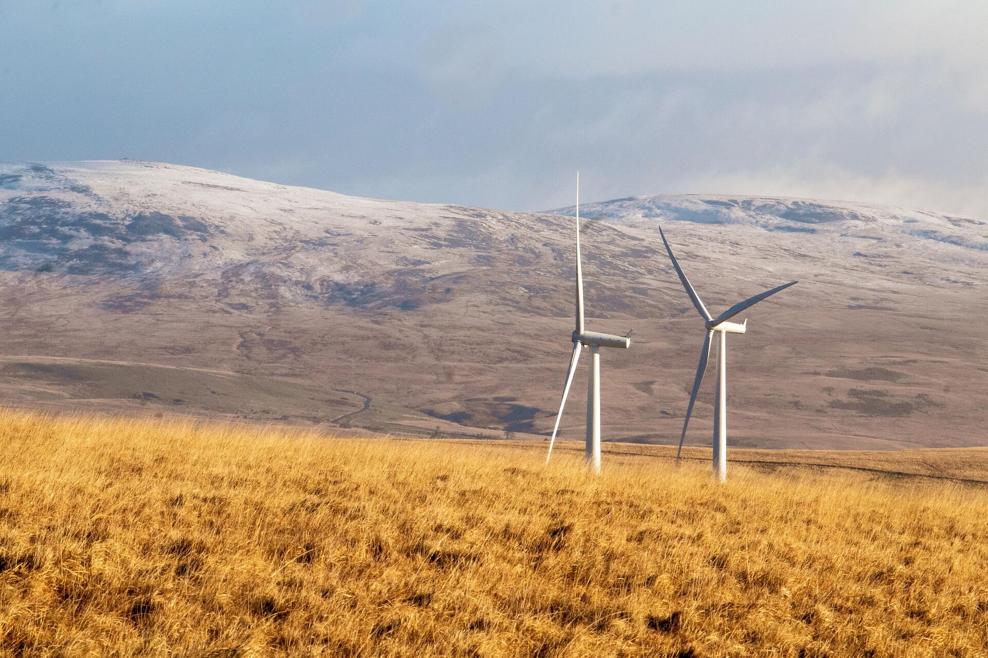 Zwei Windräder in einer hügeligen Landschaft mit Schneeresten auf den Bergen im Hintergrund.