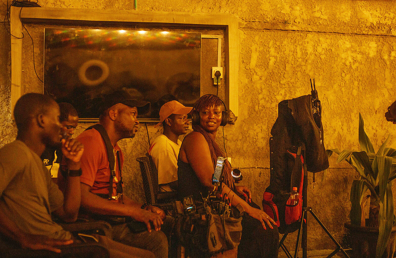Fünf Personen sitzen nebeneinander in einem Raum mit gelber Wand, einige tragen Kameraausrüstung, im Hintergrund ein großer Bildschirm.