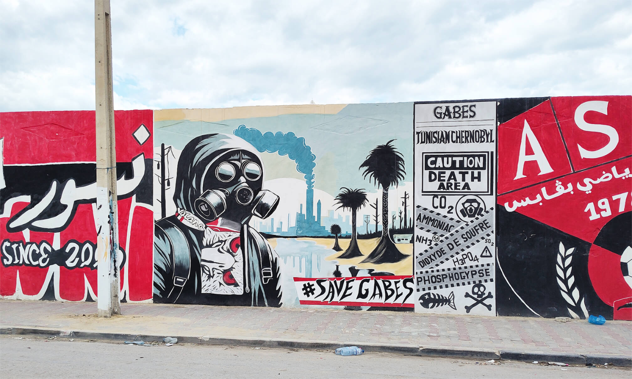 Wandbild mit Person in Gasmaske vor rauchenden Fabrikschloten, Palmen und Warnschilden mit Texten wie 'Caution Death Area' und '#Save Gabes'.