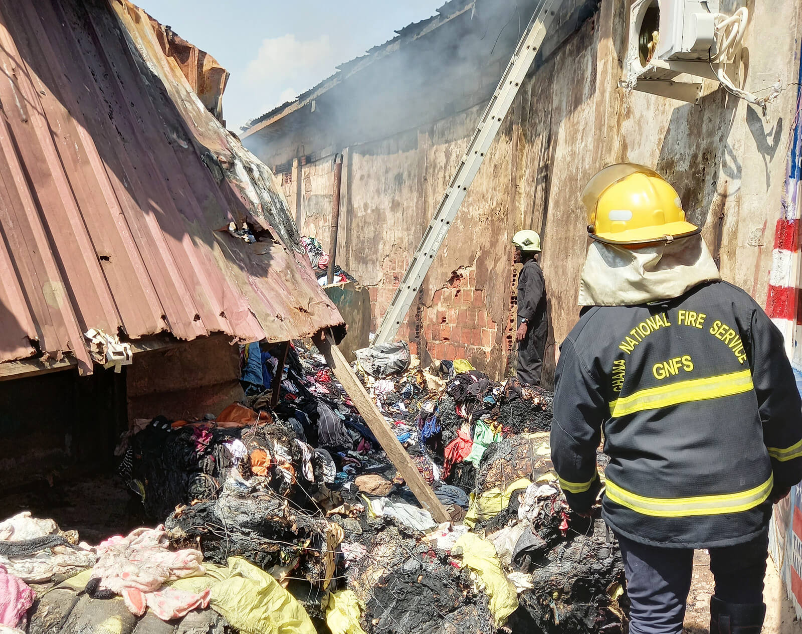 Feuerwehrleute der Ghana National Fire Service in Schutzkleidung vor einem abgebrannten Gebäude mit verkohltem Müll und Rauch.