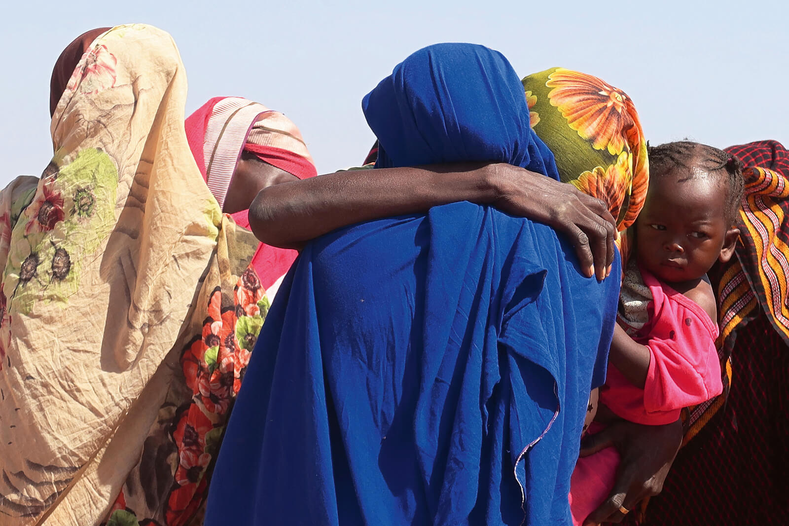 Eine Gruppe von Frauen, zwei davon in einer Umarmung, dazwischen ein kleines Kind, das eine der Frauen auf dem Arm hält.