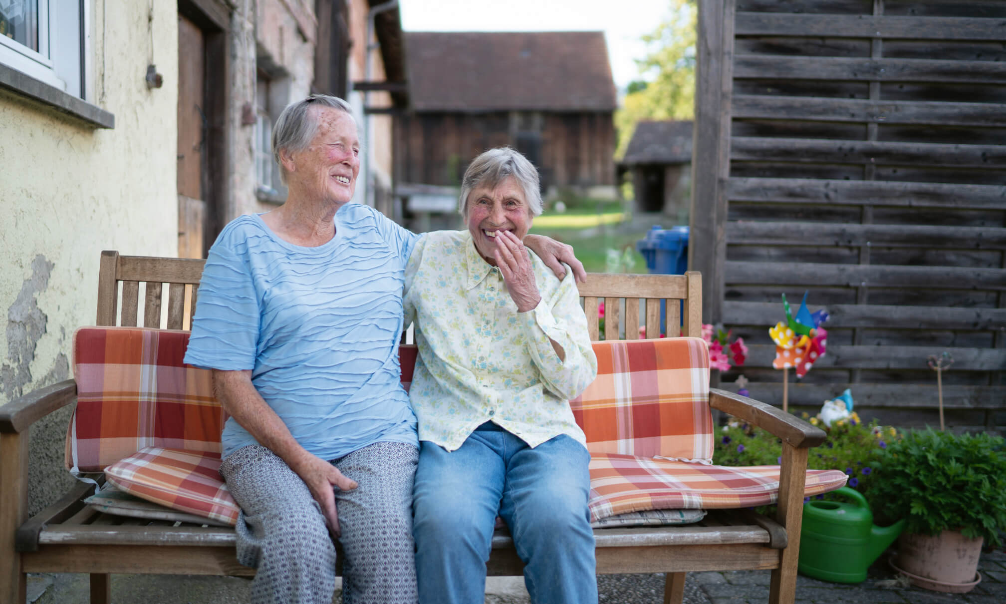 Zwei Seniorinnen sitzen lachend nebeneinander auf einer Bank unter freiem Himmel.