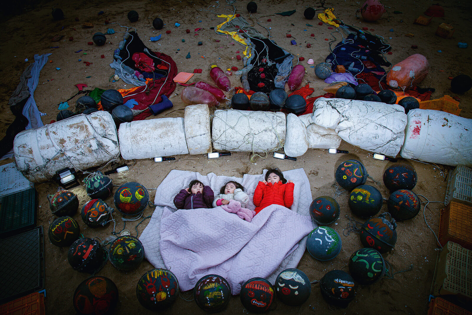 Drei Kinder schlafen am Strand, umgeben von bemaltem Müll.