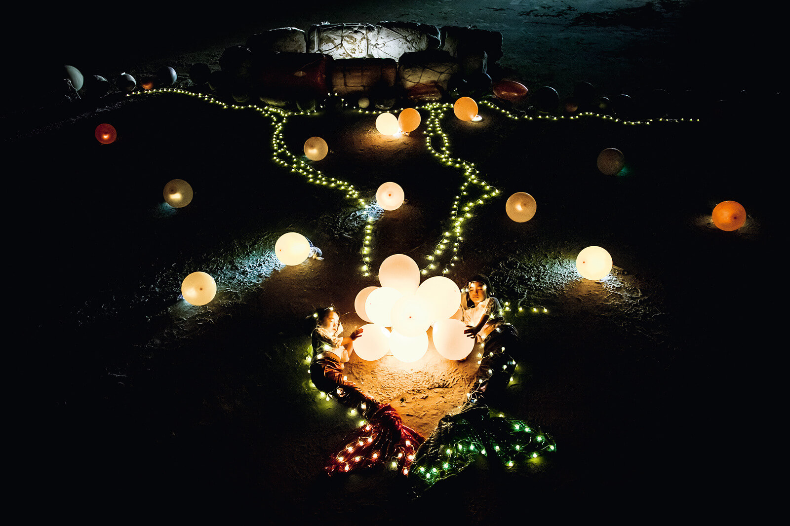 Zwei Kinder sitzen nachts zwischen leuchtenden Ballons und Lichterketten.