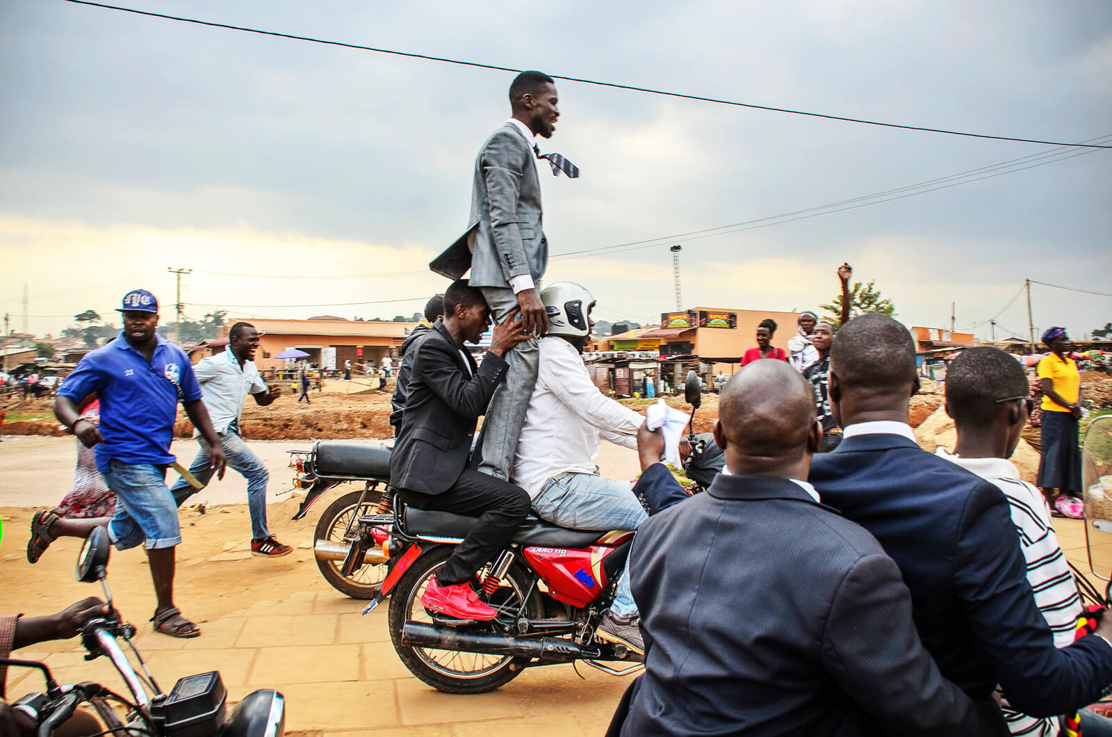 Menschenmenge um ein Motorrad, auf dem ein Mann im Anzug stehend durch eine Straße fährt.