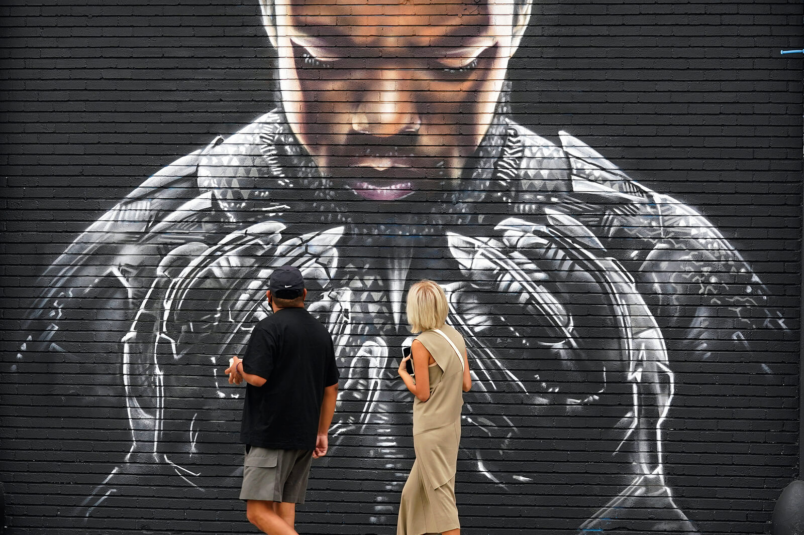Zwei Personen stehen vor einem großformatigen Wandgemälde von Black Panther auf einer schwarzen Ziegelwand.