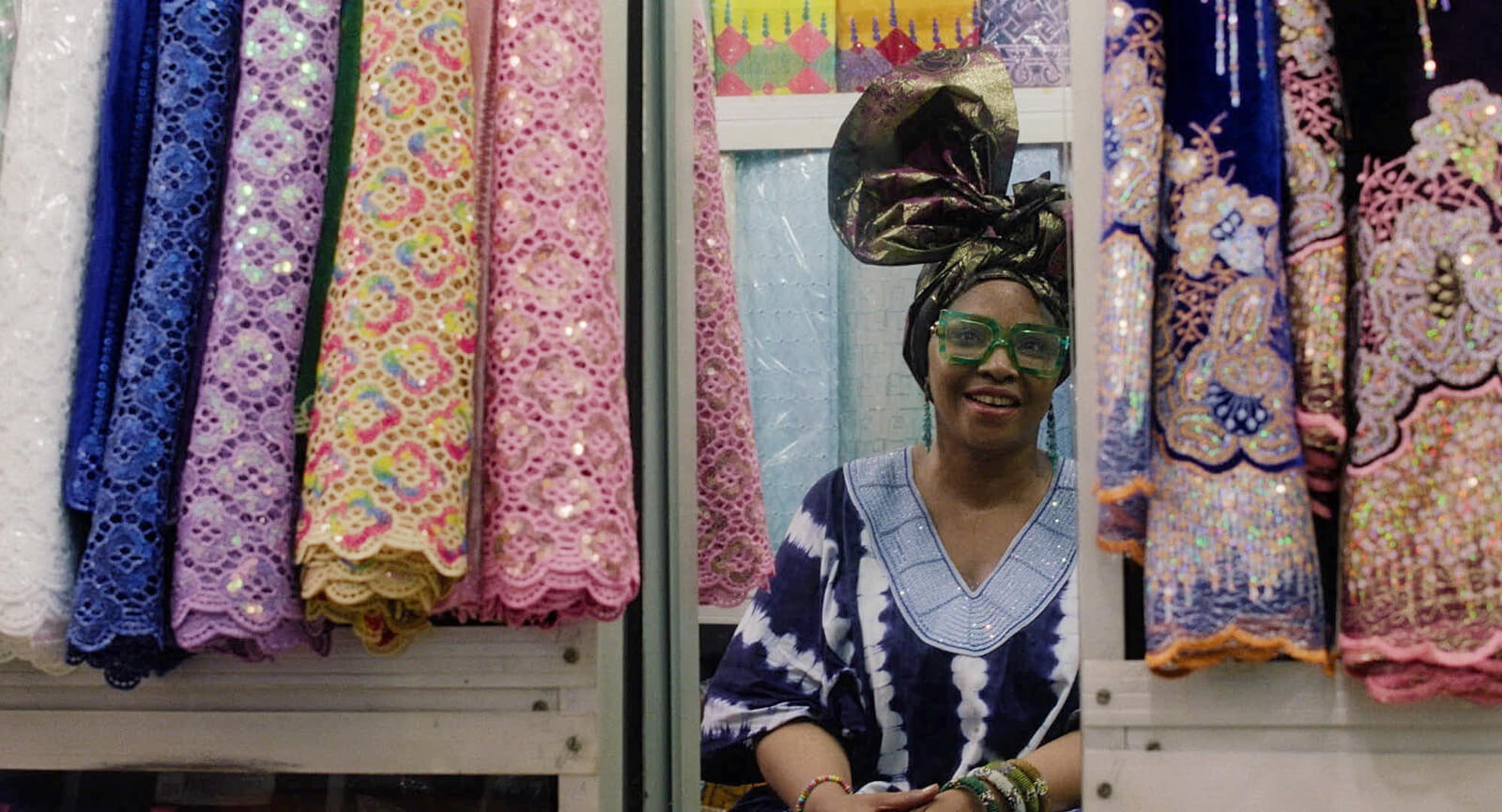 Frau mit Kopftuch und grüner Brille sitzt lächelnd zwischen bunten Stoffen und Spitzen in einem Laden.