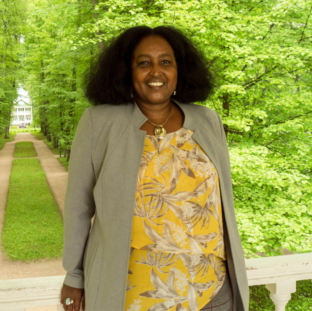 Lächelnde Amina Mire in gelber Bluse und grauem Blazer steht in einem grünen Park.